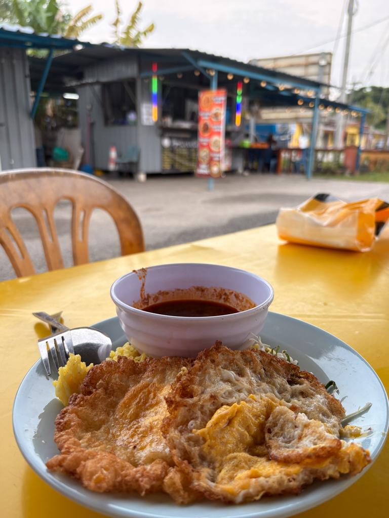 A fried egg breakfast sits atop a yellow plastic table with a metal food stall in the background.